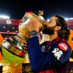 RCB captain Rajat Patidar kissing the IPL 2026 trophy under stadium floodlights, with golden confetti and the charged atmosphere of a championship night in the background.