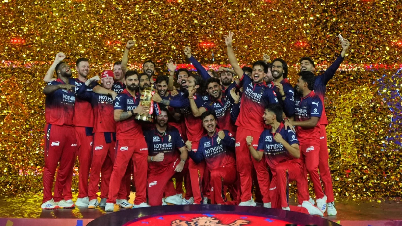 RCB players celebrating their IPL 2026 championship title on stage, holding the winners' trophy amidst a shower of golden confetti, with the Royal Challengers Bengaluru Champions 2026 podium in the foreground.